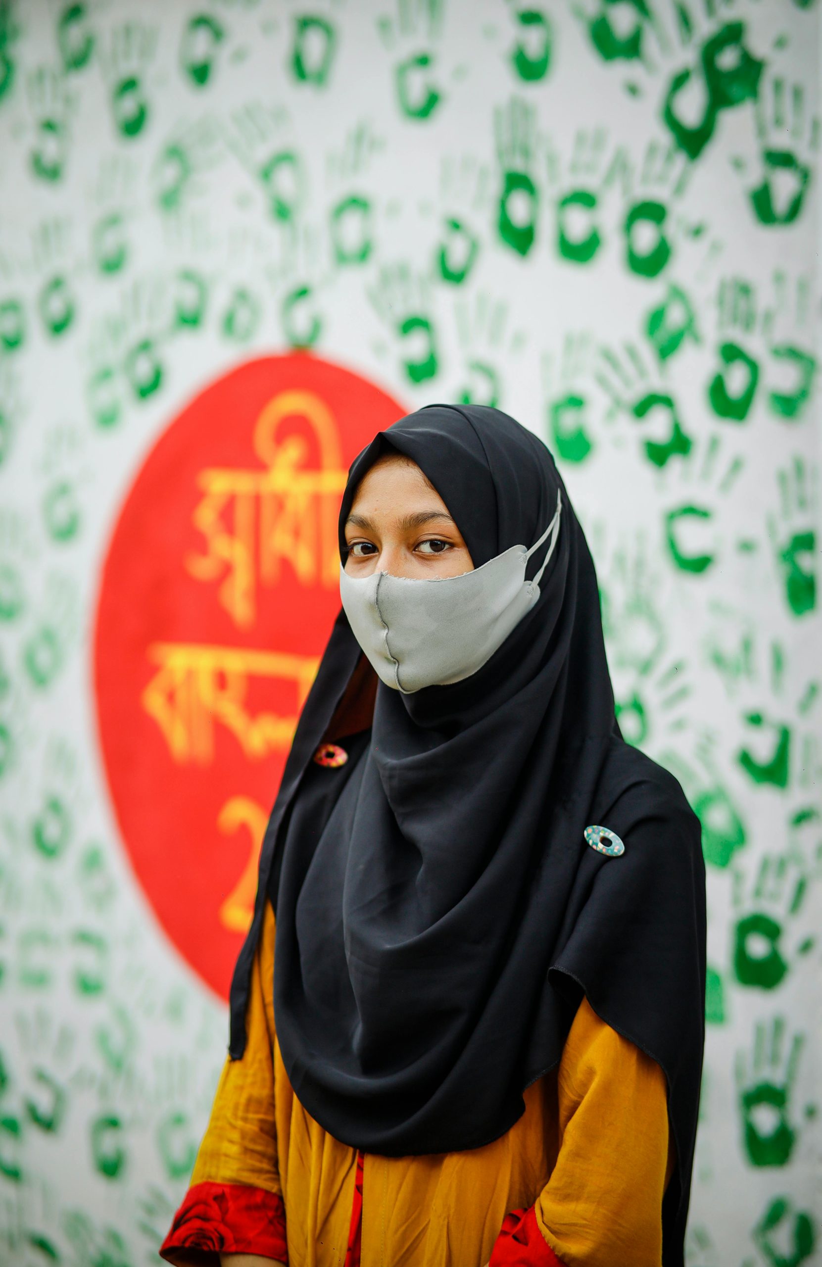 Portrait of a young woman wearing a hijab and mask against a colorful backdrop in Bangladesh.