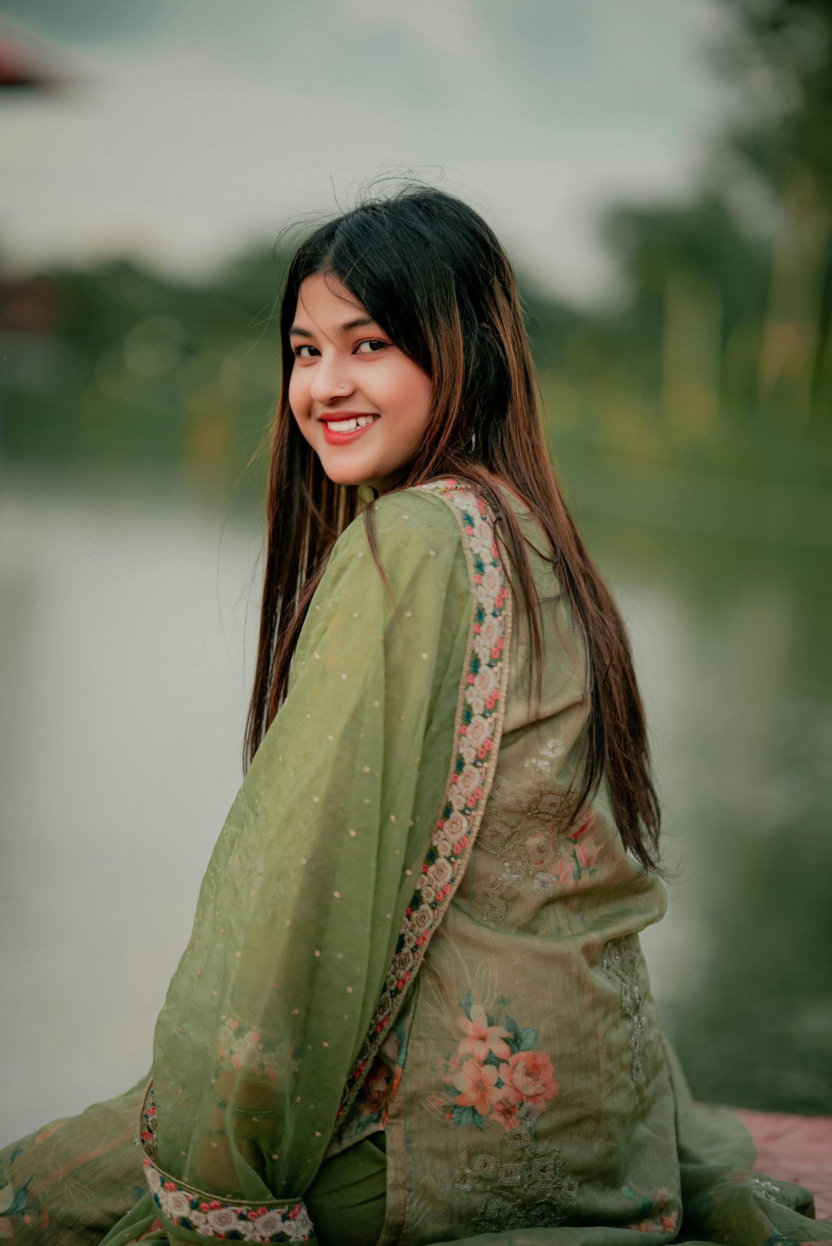 A smiling woman in traditional green attire with floral embroidery by a serene lake in Bangladesh.