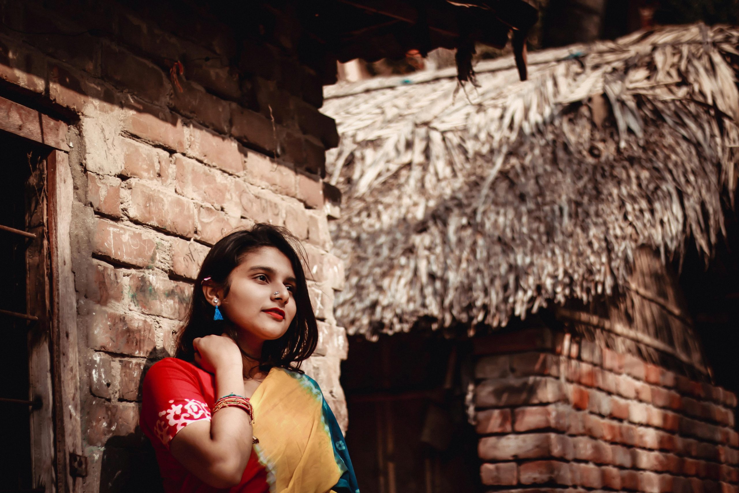 Charming ethnic female in national earring and piercing wearing colorful dress looking away near shabby old building